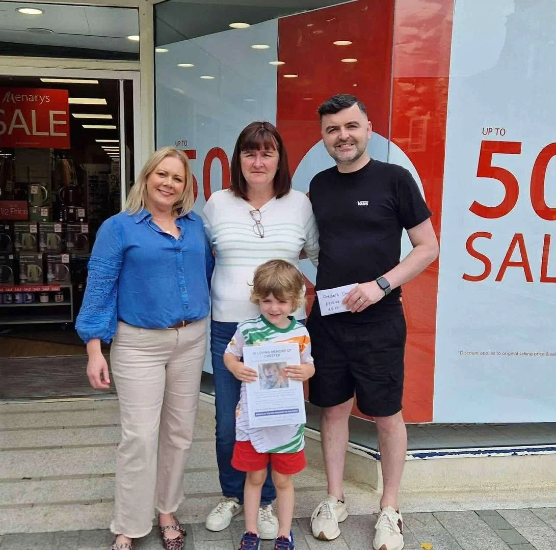 A group of five people, including a woman, a man, a young boy, and two other women, standing outside a store with sale signs. The young boy is holding a printed document.