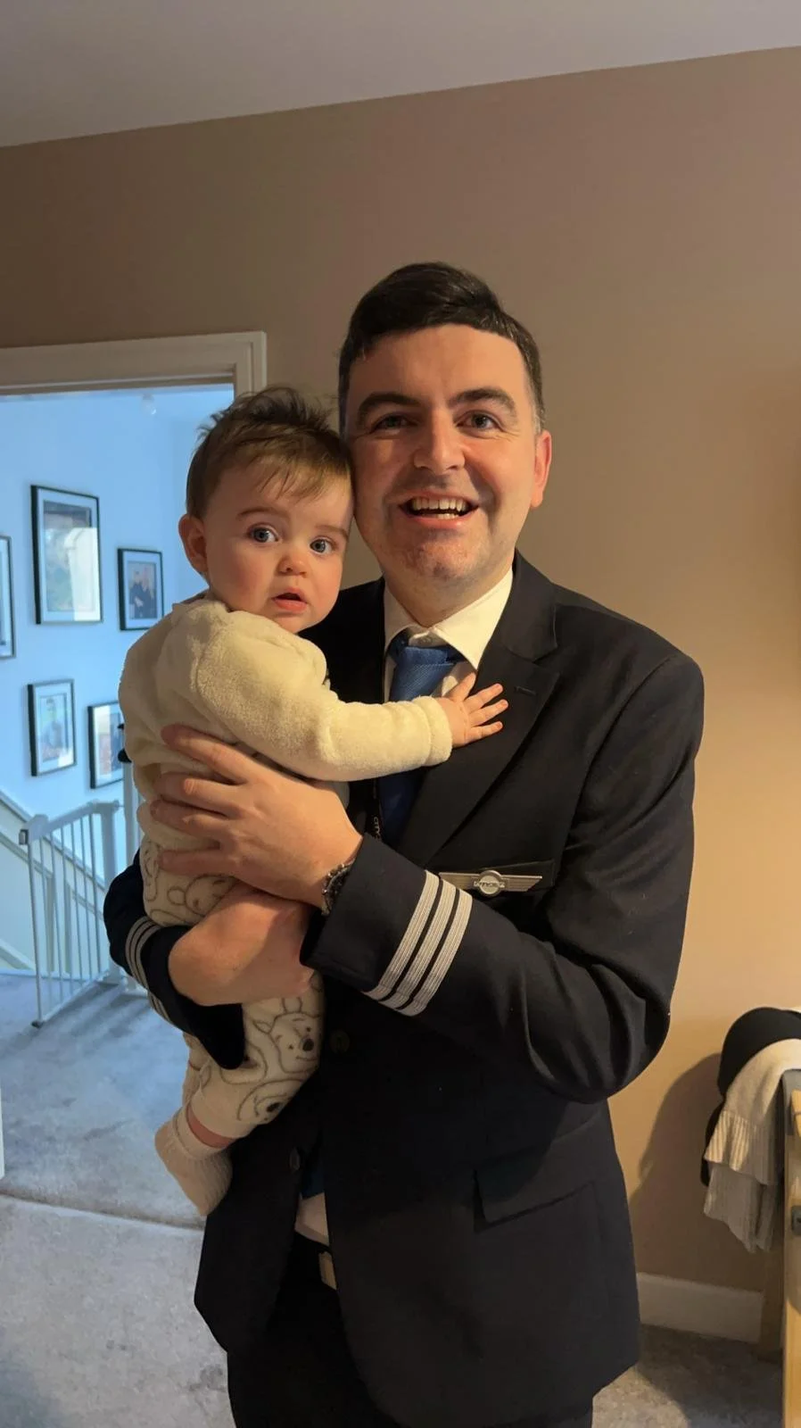 A man in a pilot uniform holding a young child inside a house, with framed pictures on the wall in the background.