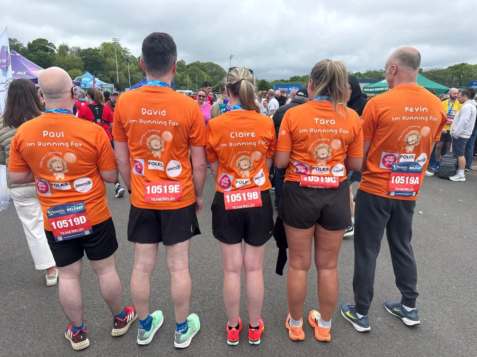 Group of five runners wearing orange shirts at a race event, standing with their backs to the camera. Each shirt has the person's name and 'I'm Running For' printed on it, along with sponsored patches and race bibs.