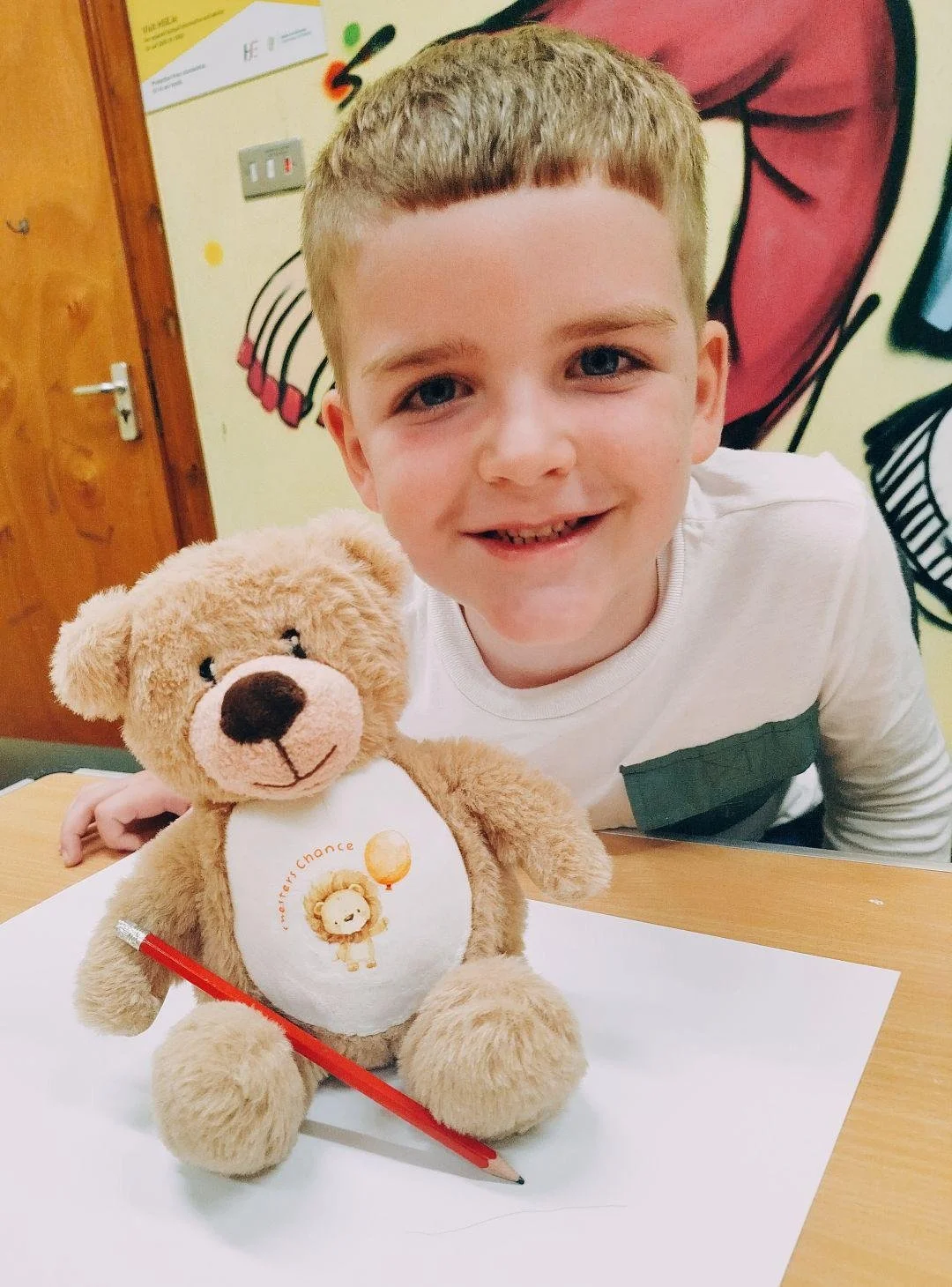 A young boy with short light brown hair and a white shirt smiling at the camera, sitting at a desk with a stuffed teddy bear on a sheet of paper and a red pencil. The teddy bear has a white stomach with an illustrated lion holding a balloon and Frenc