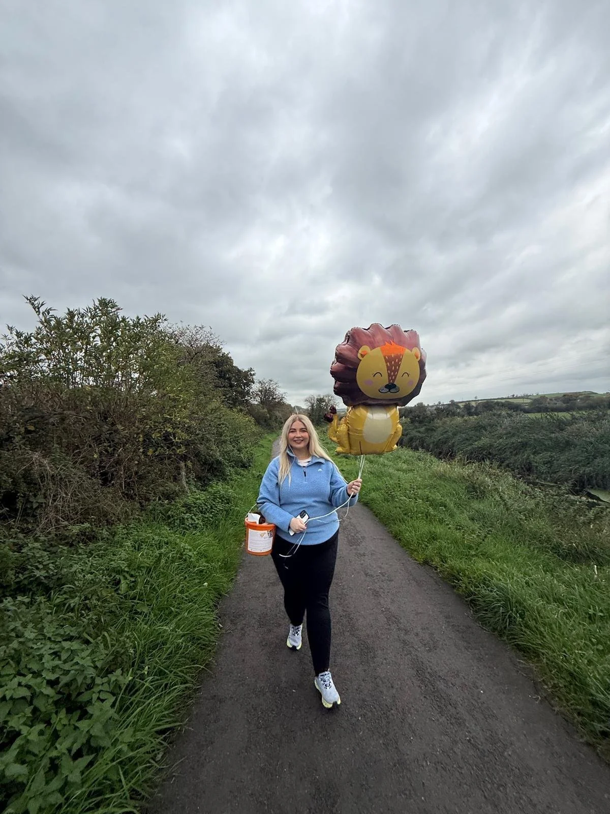A young woman in a blue jacket walking on a narrow rural path with green bushes on either side, holding a lion-shaped helium balloon and a small orange bucket, under a cloudy sky.