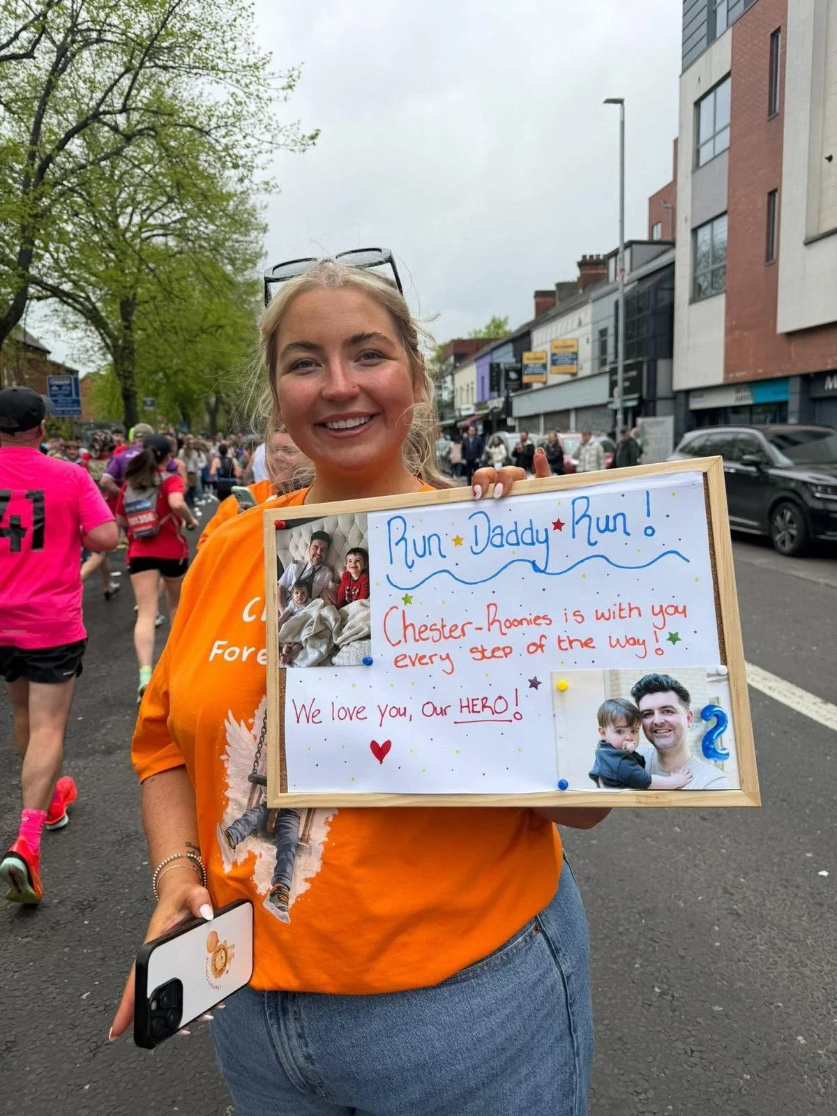 A woman at a marathon is holding a sign with photos and messages, surrounded by runners and spectators on a city street with trees and buildings.