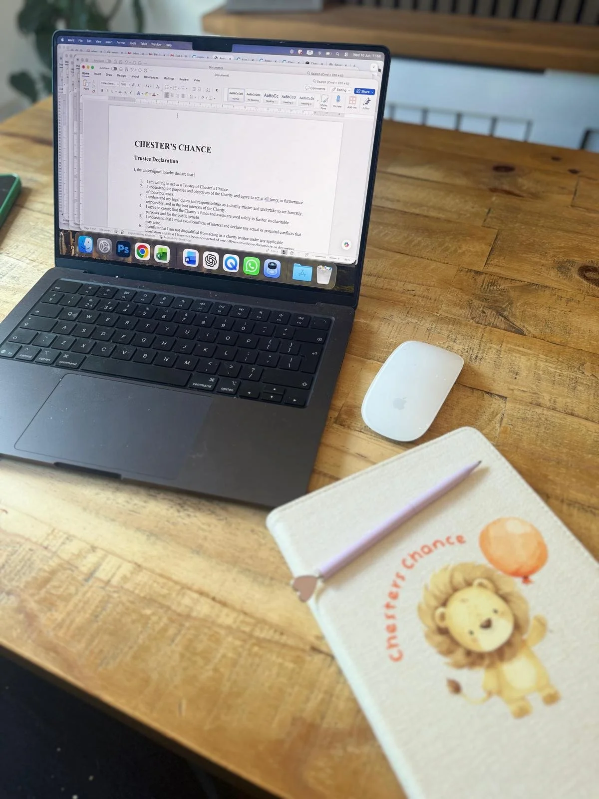 A laptop on a wooden desk displaying a document titled "Chester’s Chance" with the text of a trustee declaration, a white mouse, a notebook with a pen, and a birthday card with a lion illustration.