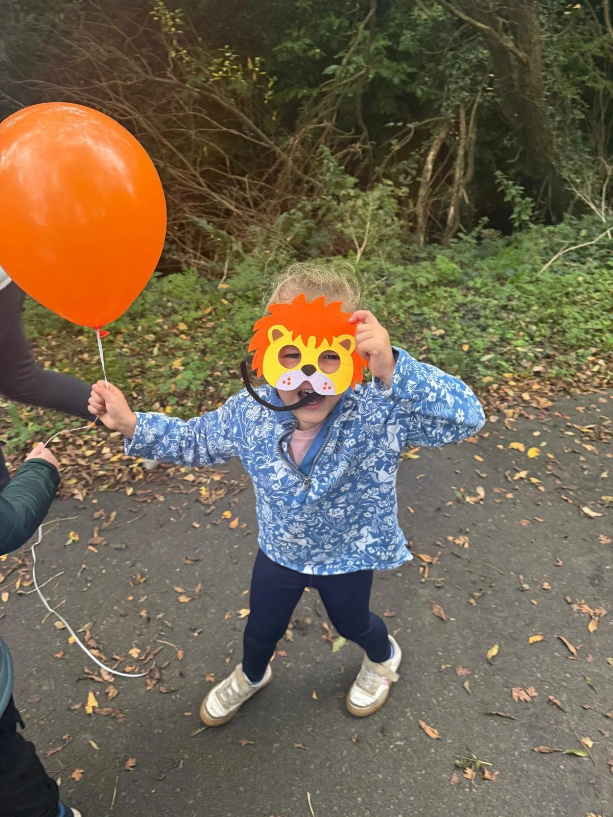A young child outdoors on a sidewalk, wearing a lion mask and holding an orange balloon.