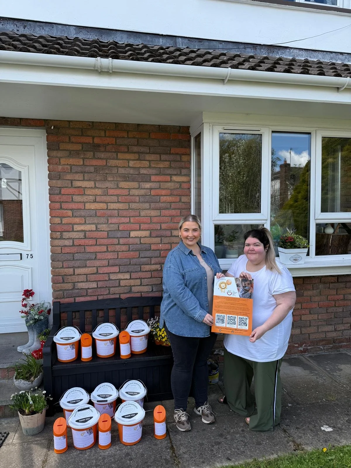 Two women standing outside a brick house with a window and door, holding a flyer, and smiling. On a bench next to them are donation buckets, flyers, and potted plants.