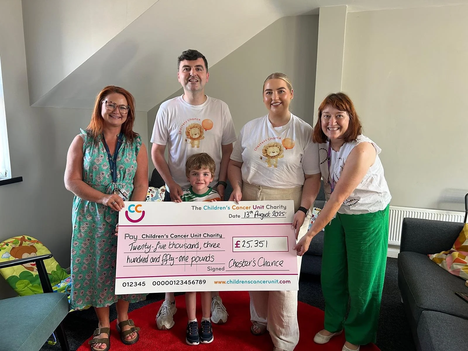 Group of six people, including a young boy, holding a large ceremonial check for £25,351 made out to the Children's Cancer Unit Charity, in a room with chairs and a window.