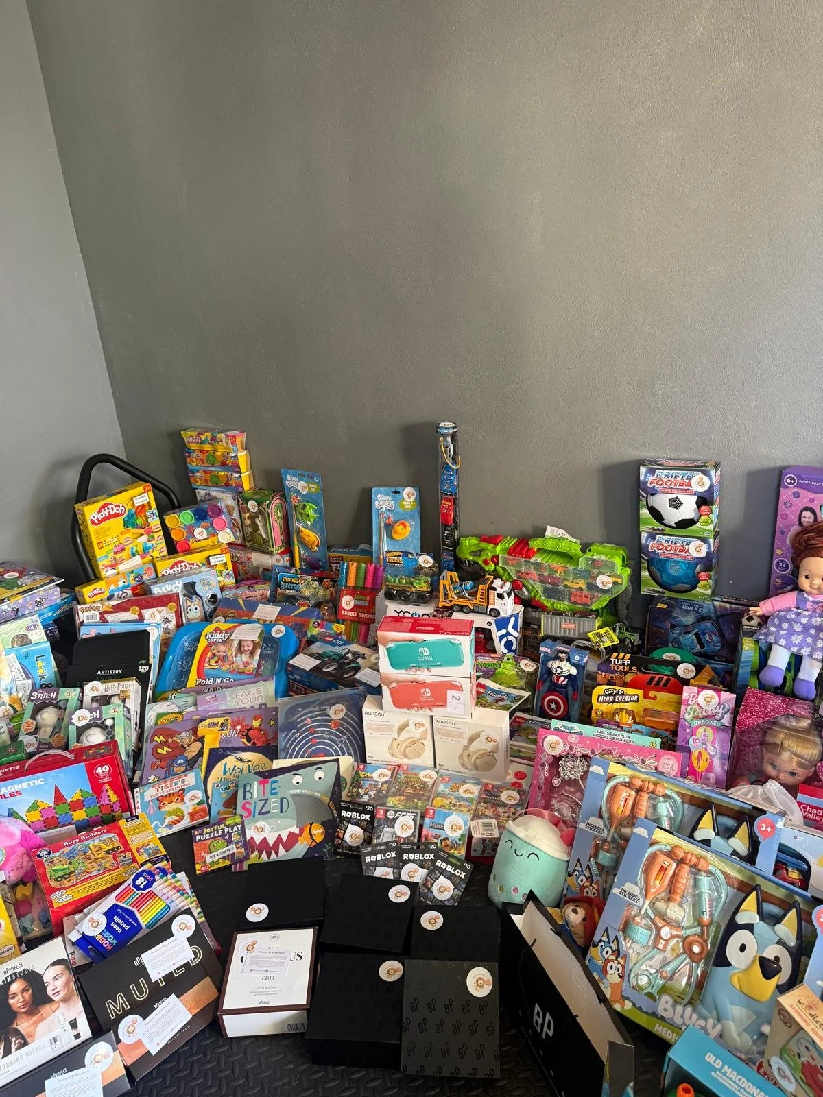 A large collection of toys, including dolls, action figures, building blocks, and electronic gadgets, arranged on a table against a plain gray wall.
