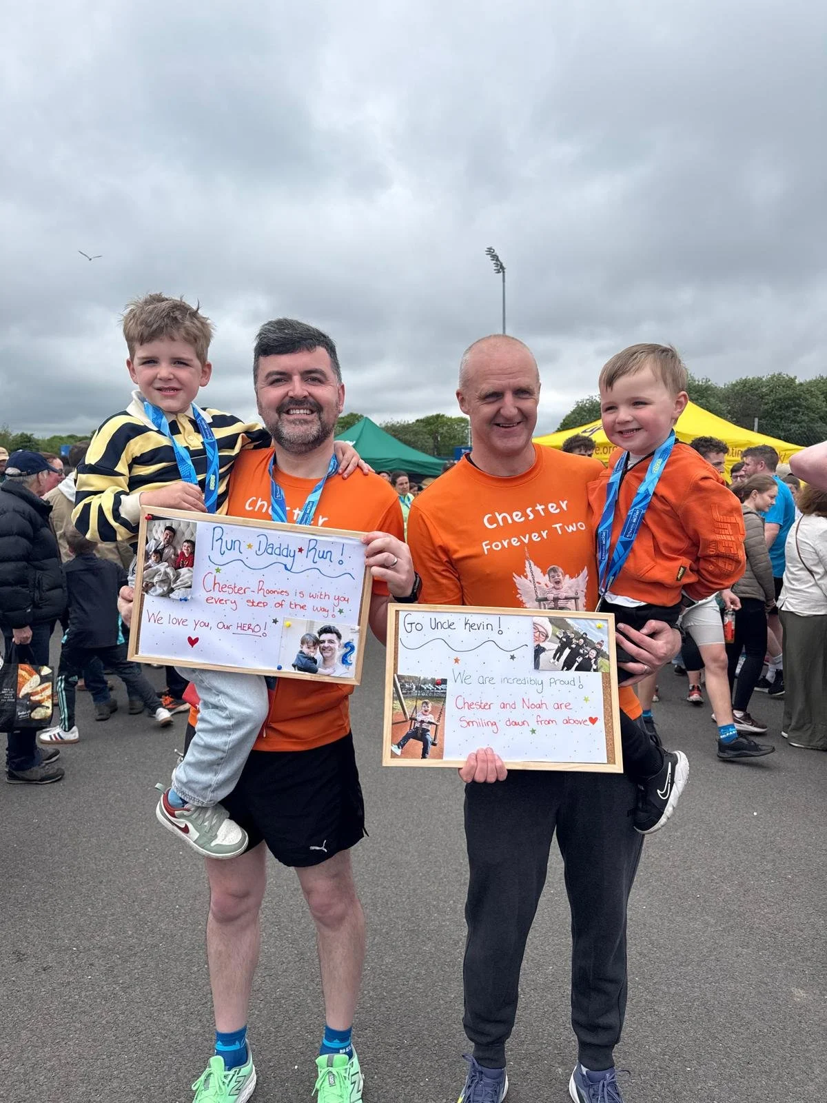 Two men holding children at a charity event with signs supporting children. One man and child are wearing orange shirts and medals.