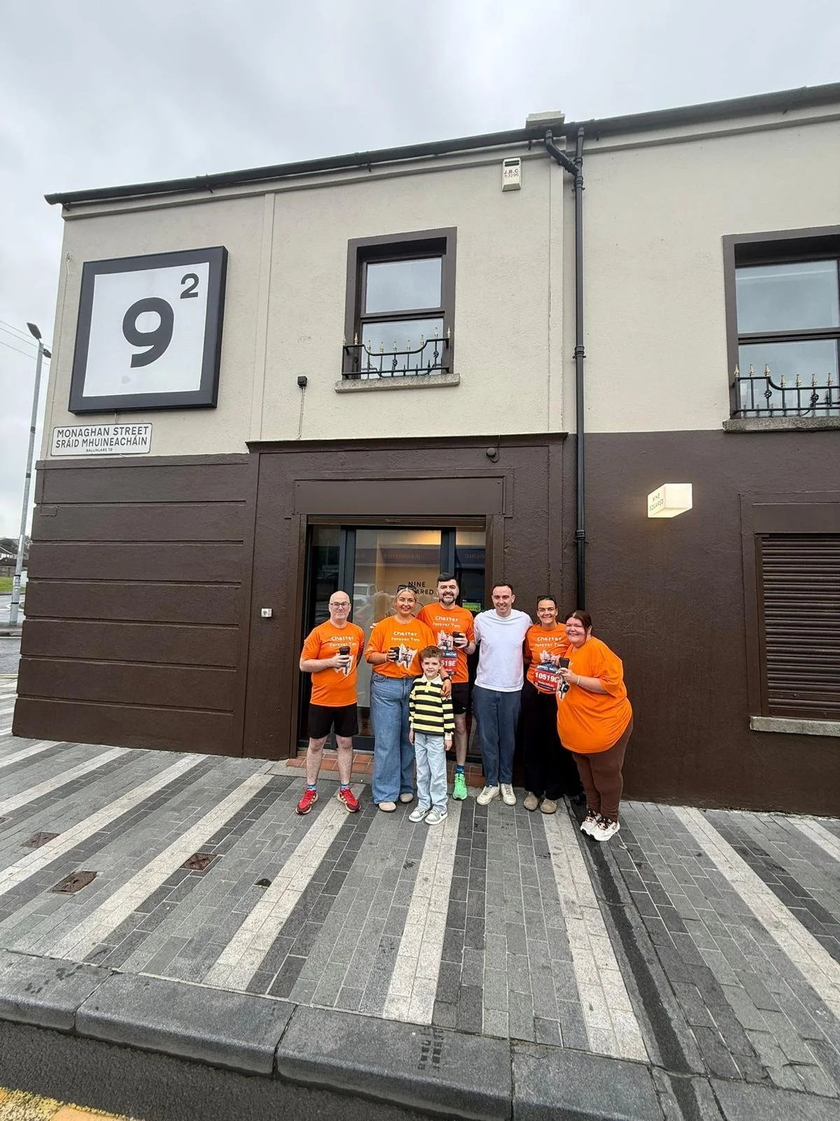 Group of six adults and one child standing outside a building on a sidewalk, some adults wearing orange Shirts and holding flyers, smiling at the camera.