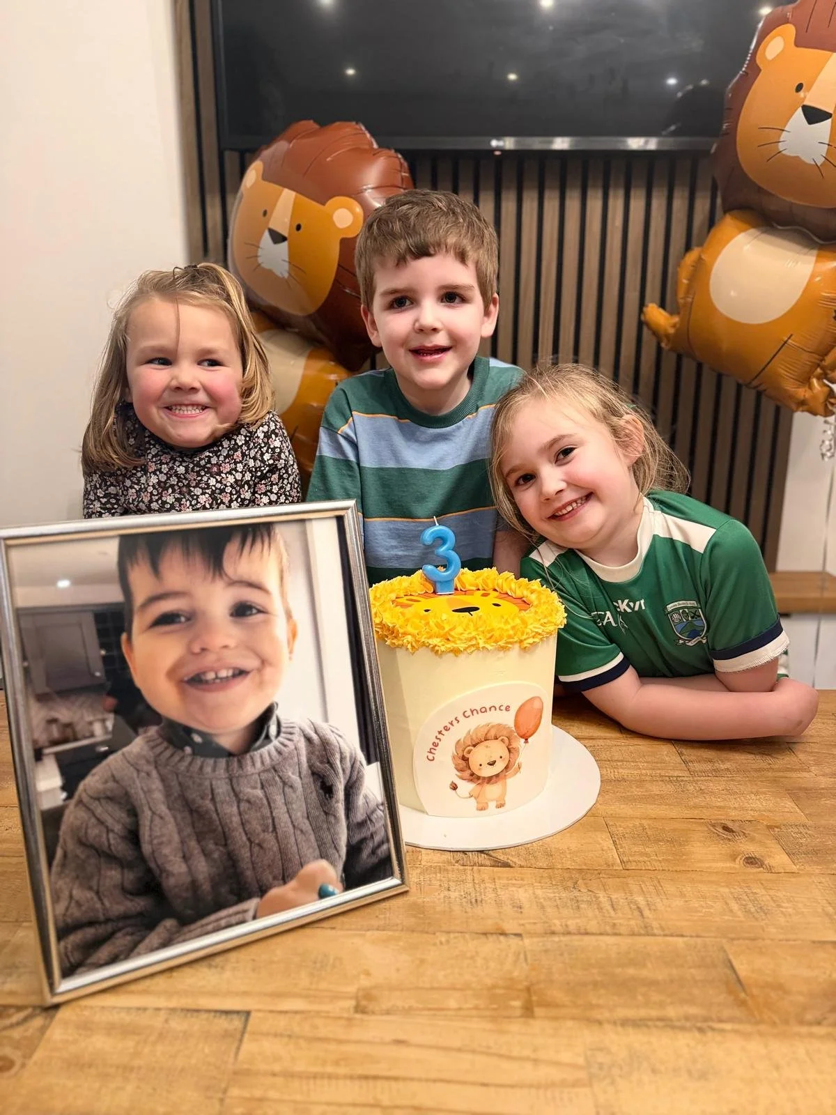 Three children celebrating a third birthday, with a birthday cake decorated with a lion face, a picture of a younger boy, and lion-themed balloons in the background.