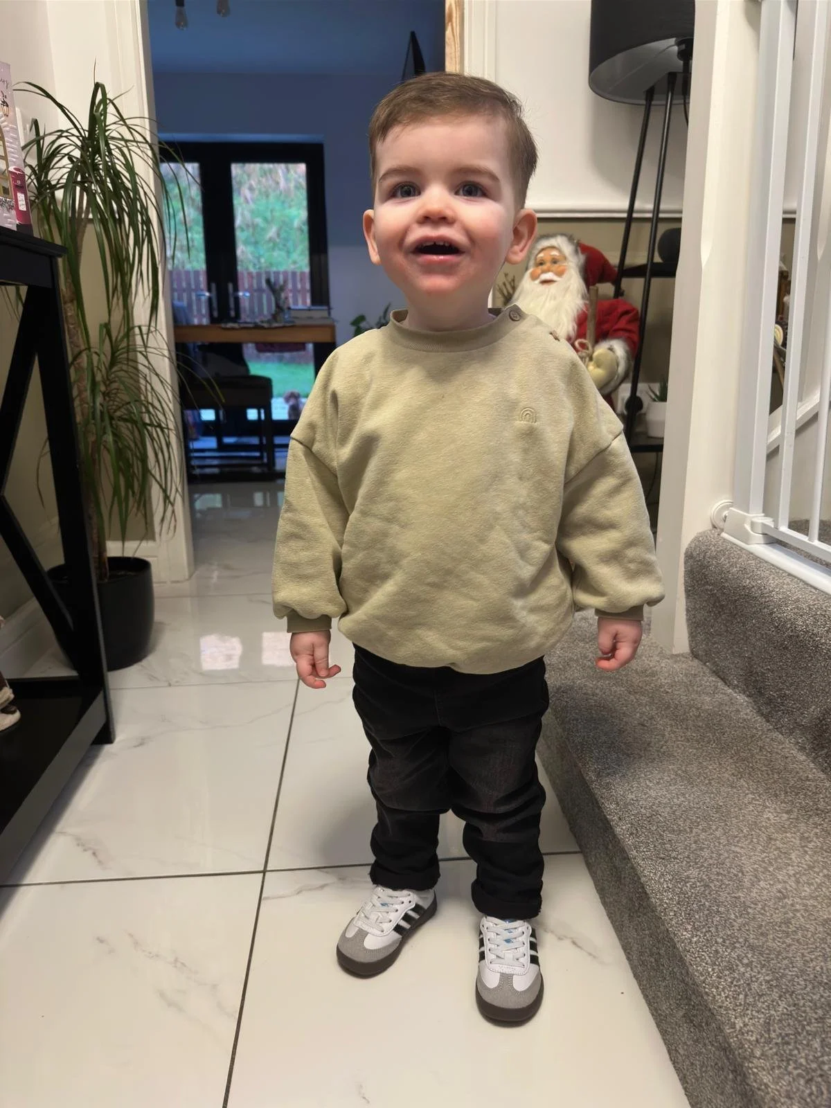 A young boy with short light brown hair and blue eyes standing indoors. He is wearing a beige sweatshirt, black pants, and gray sneakers with white laces. He has a cheerful expression. In the background, there is a Christmas decoration of Santa Claus, a potted plant, and a view of the backyard through glass doors.