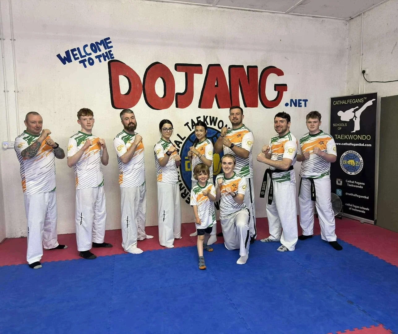 Group of ten taekwondo practitioners, including children, posing with fists up in a taekwondo studio with a large 'Welcome to the Dojang.net' sign on the wall and a banner for Cathal Fegan Taekwondo Schools.