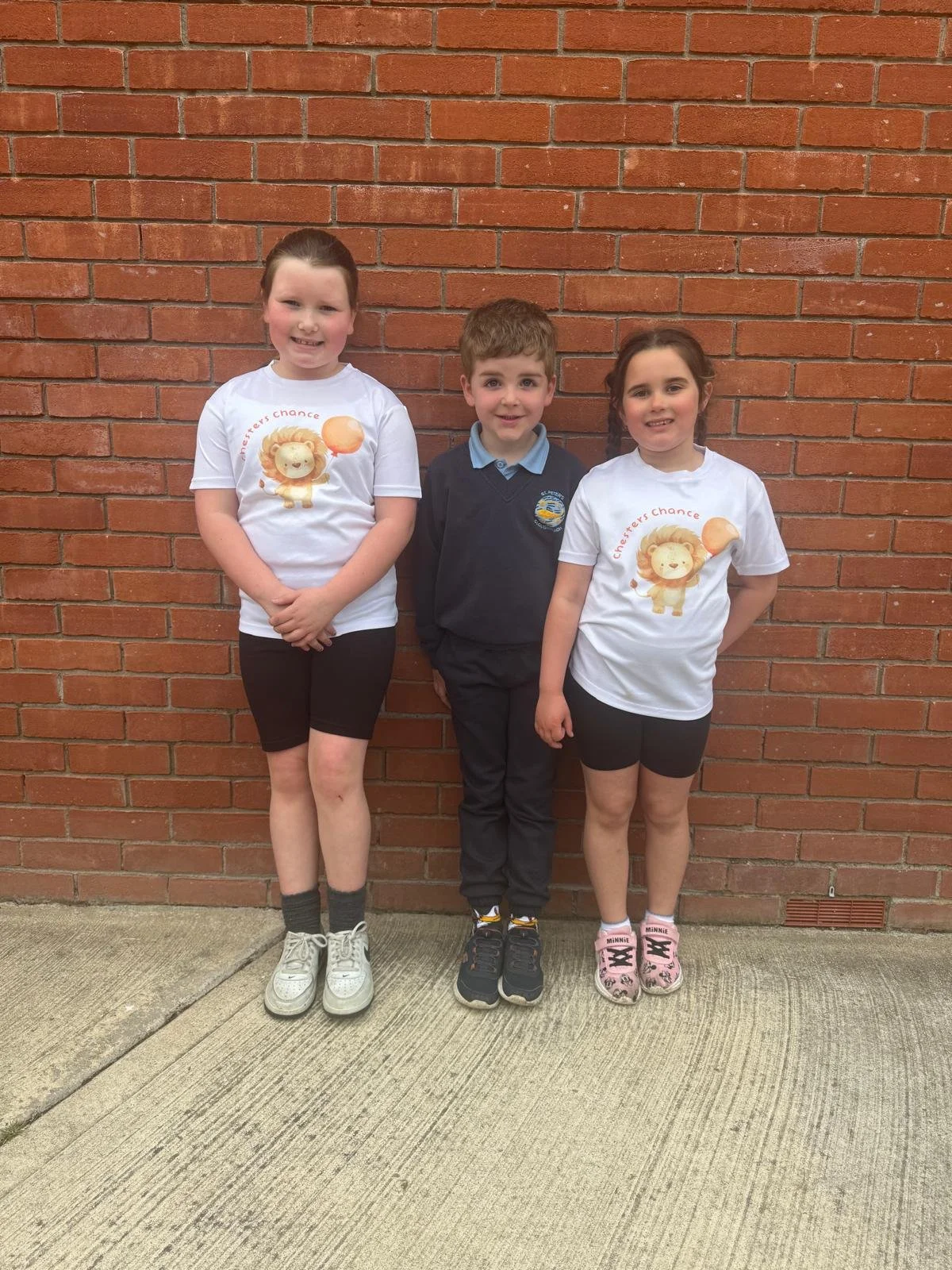 Two young girls and one young boy standing in front of a red brick wall. The girls are wearing matching white t-shirts with a lion and a speech bubble, and black shorts. The boy is in a dark uniform with a light blue collar. All three are smiling.