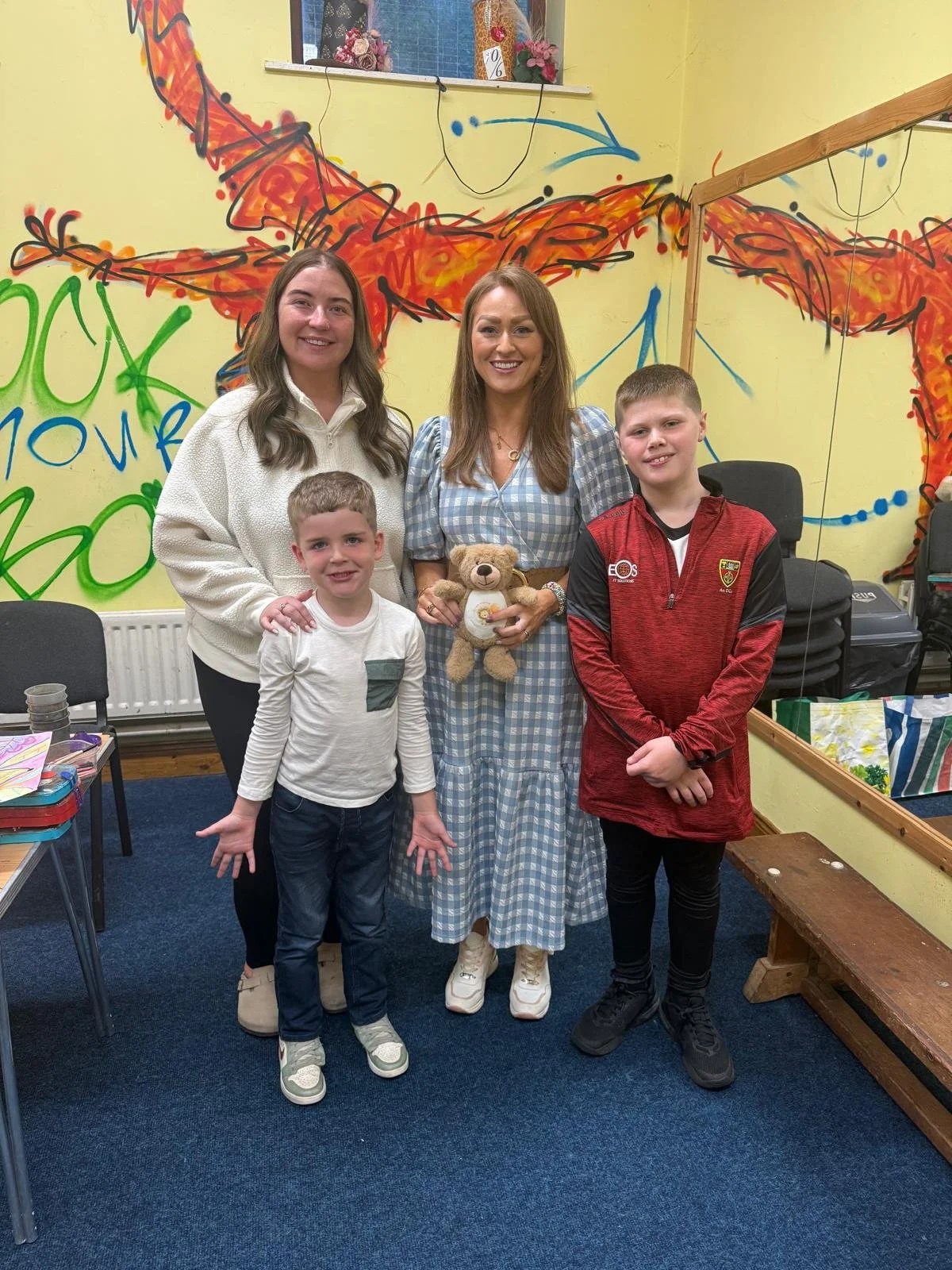 Five people, two women and three boys, standing together in a room with colorful graffiti-style art on the wall behind them. The woman in the middle is holding a teddy bear. They are smiling at the camera.
