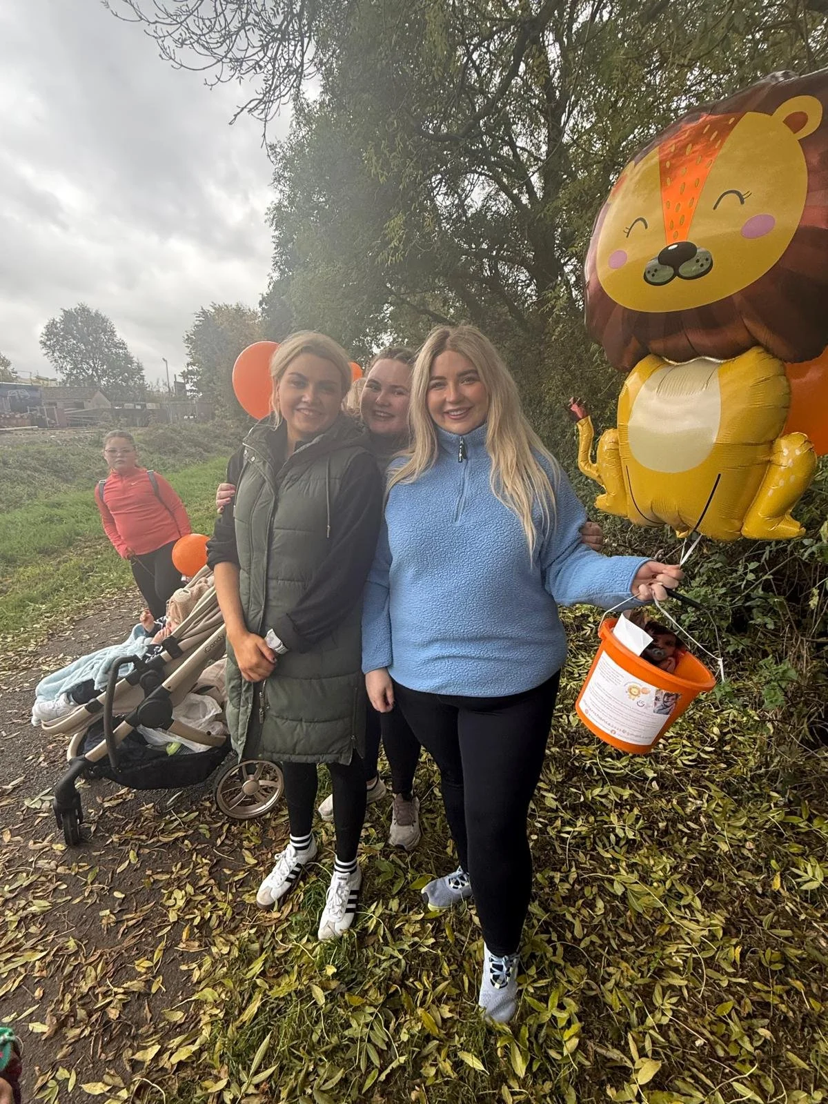 Group of smiling women walking outdoors on a cloudy day, holding orange themed balloons and bucket, with children and a stroller in the background.