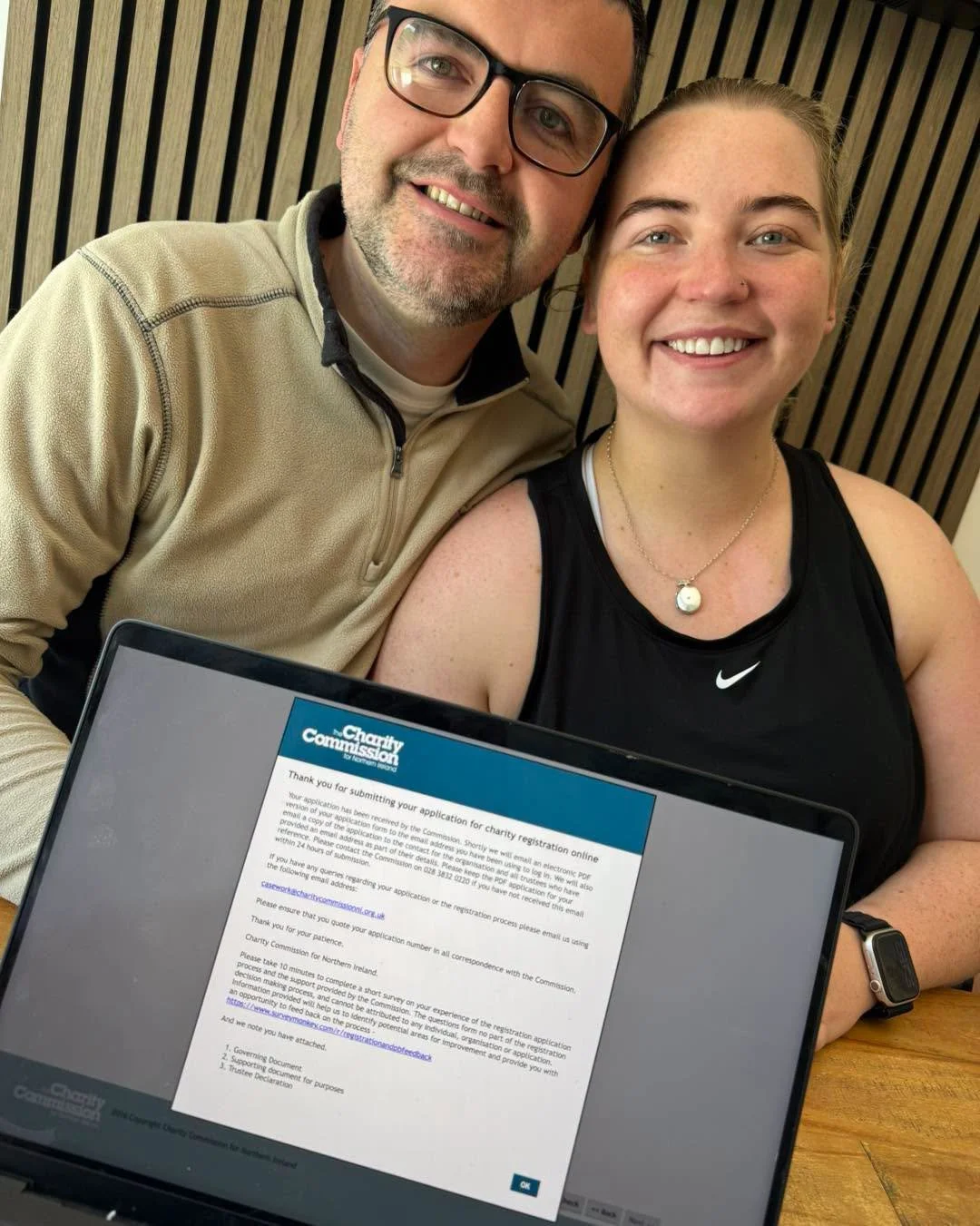 A smiling man and woman taking a selfie together, with a laptop displaying a charity registration confirmation on the table in front of them.