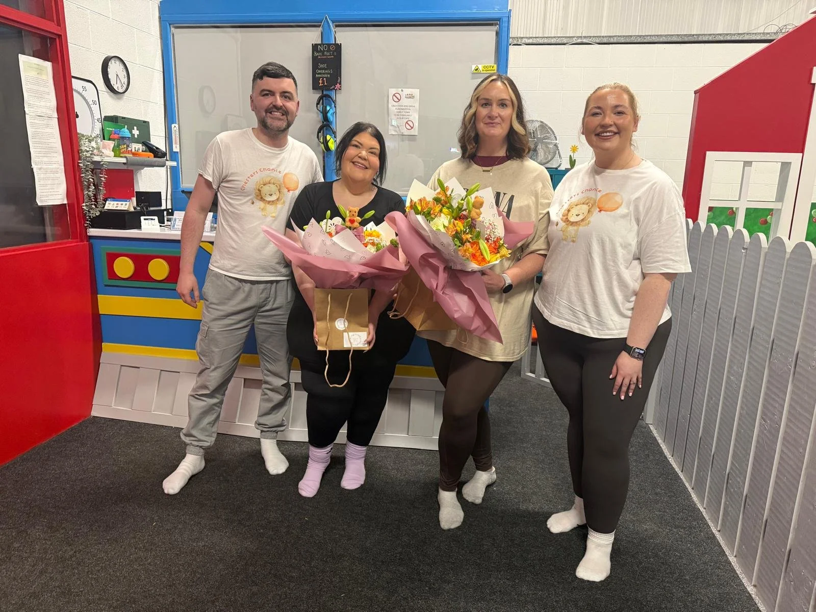 Four smiling adults standing together indoors, three women and one man, holding bouquets of flowers and a gift bag, all wearing socks.