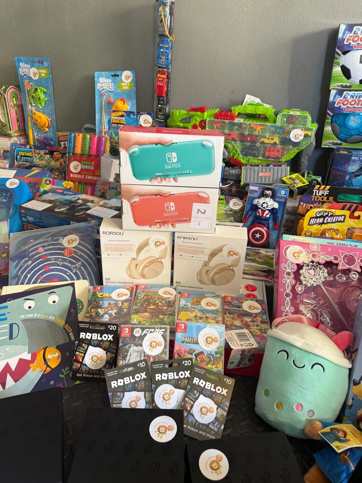 A variety of toys and games displayed on a table, including Nintendo Switch consoles, wireless headphones, Roblox gift cards, figurines, and stuffed toys, with some larger toy sets and packaging visible in the background.