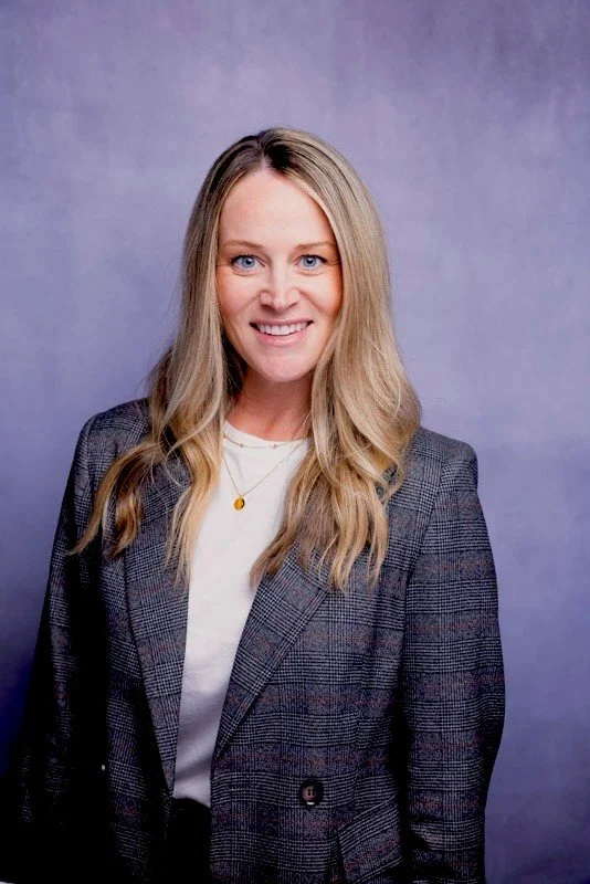 Founder and non-profit executive with long blonde hair, wearing a gray plaid blazer and a white top, smiling at the camera against a purple background.
