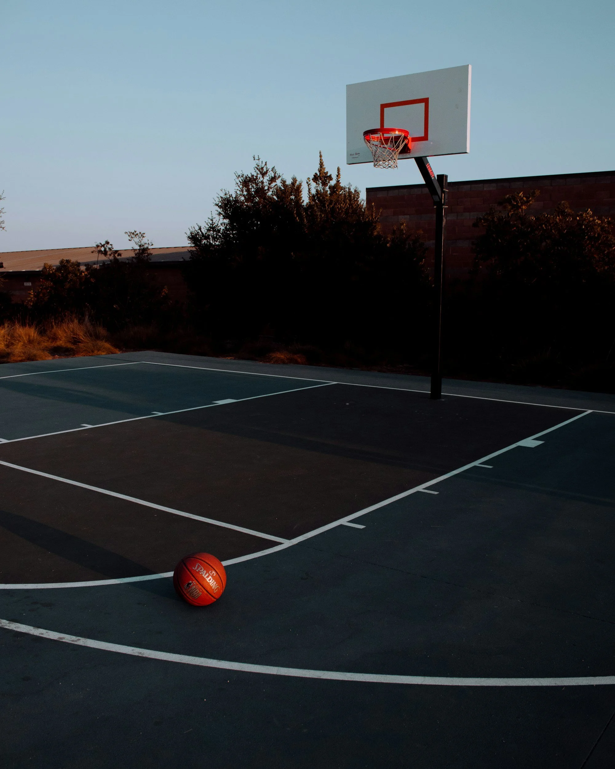 An empty outdoor basketball court at dusk with two basketballs on the court and a basketball hoop in the background.