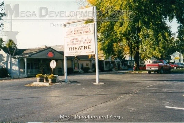An empty parking lot with a large sign featuring businesses There are trees and parked cars in the background.