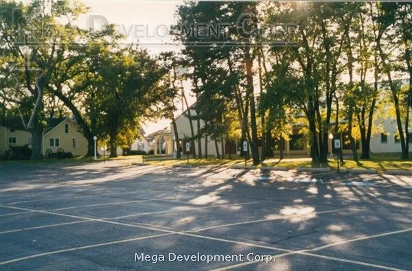 Empty parking lot with trees and a white building in the background, sunlight filtering through the trees.