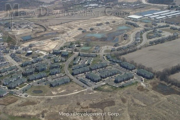 Aerial view of a residential neighborhood with houses, roads, and a park with ponds, surrounded by farmland and undeveloped land.