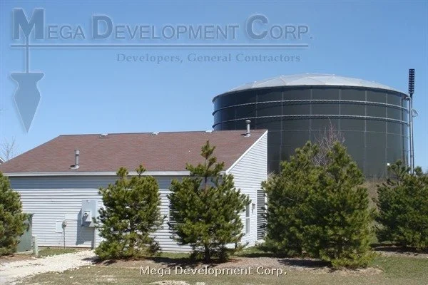 A white building with a brown roof surrounded by green trees in front of a large, black, cylindrical water tank against a clear blue sky.