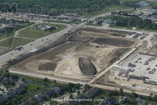 Aerial view of a construction site with cleared land, dirt piles, and construction equipment, surrounded by roads, parking lots, and residential houses.
