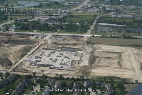 Aerial view of a construction site with cleared land and building foundations, surrounded by residential neighborhoods and green spaces.