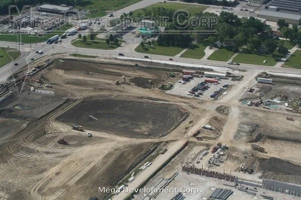 Construction site with cleared land, dirt, and construction vehicles, next to an area with parked cars, roads, and green trees.