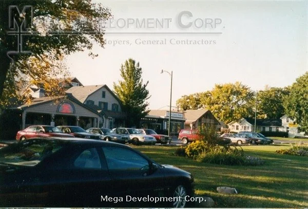 A street view featuring a row of parked cars in front of local businesses and homes, with trees and clear skies in the background.