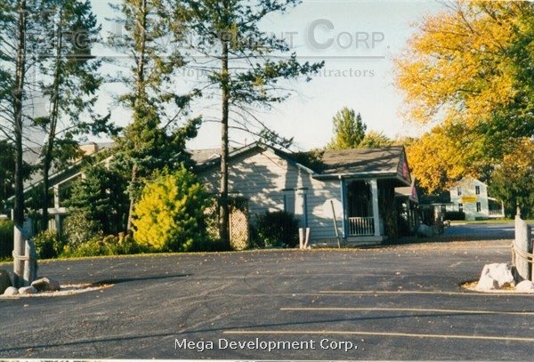 A small building with a porch, surrounded by trees, with a paved parking lot in front and a clear sky.
