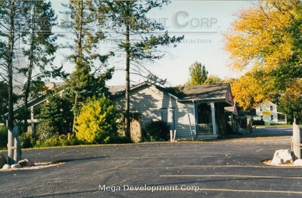 A small building with a porch surrounded by trees evolving into fall colors, with an asphalt parking lot in the foreground and white railings on either side.