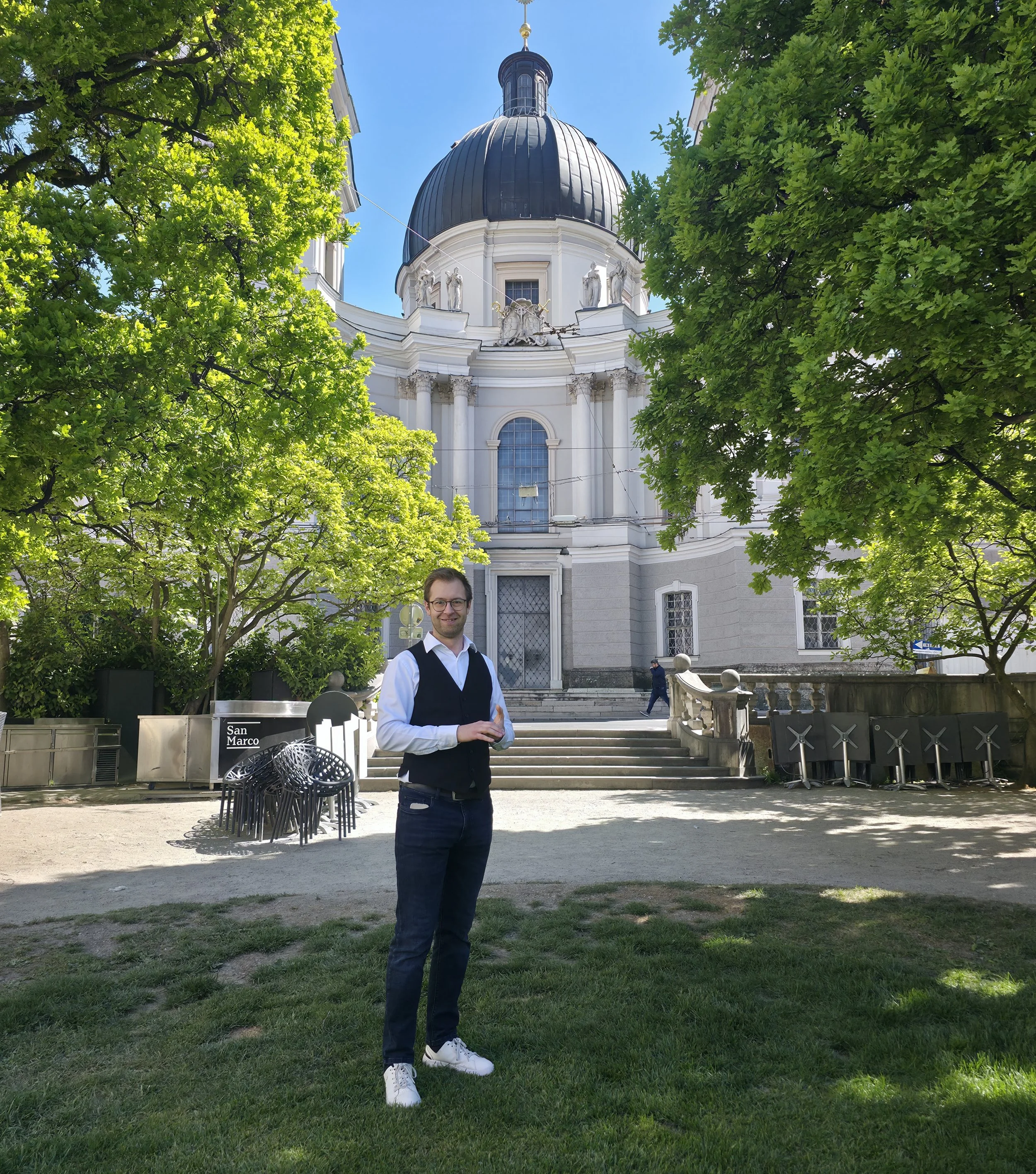 Simon steht in formeller Kleidung mit Brille steht auf einer Wiese vor einer barocken Kirche mit großen Statuen auf dem Dach und einem schwarzen, markanten Kuppeldach, umgeben von grünen Bäumen, bei sonnigem Himmel.