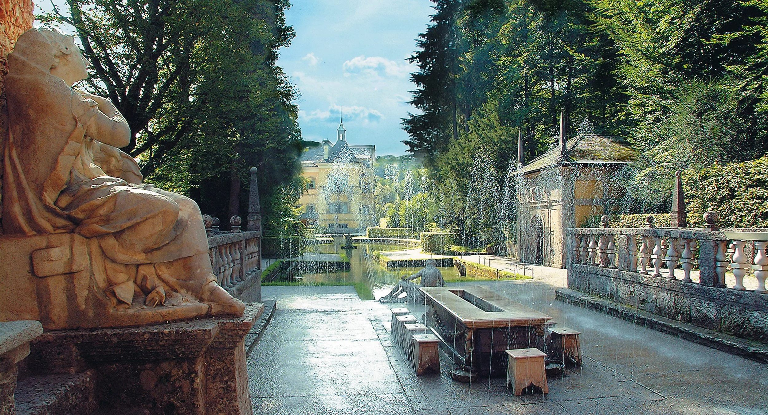 Ein gepflegter Garten mit Brunnen, Springbrunnen und Statuen, umgeben von Bäumen und einem Wasserlauf, im Hintergrund ein Schloss.