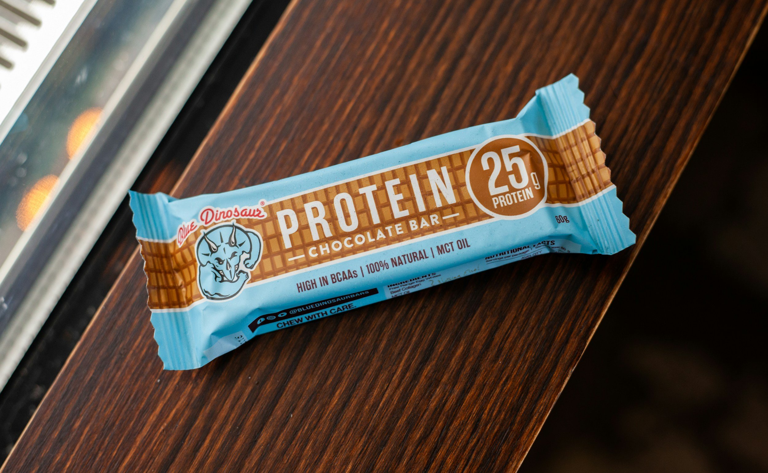 Protein chocolate bar in blue packaging with tiger logo, placed on a wooden surface near a glass window.