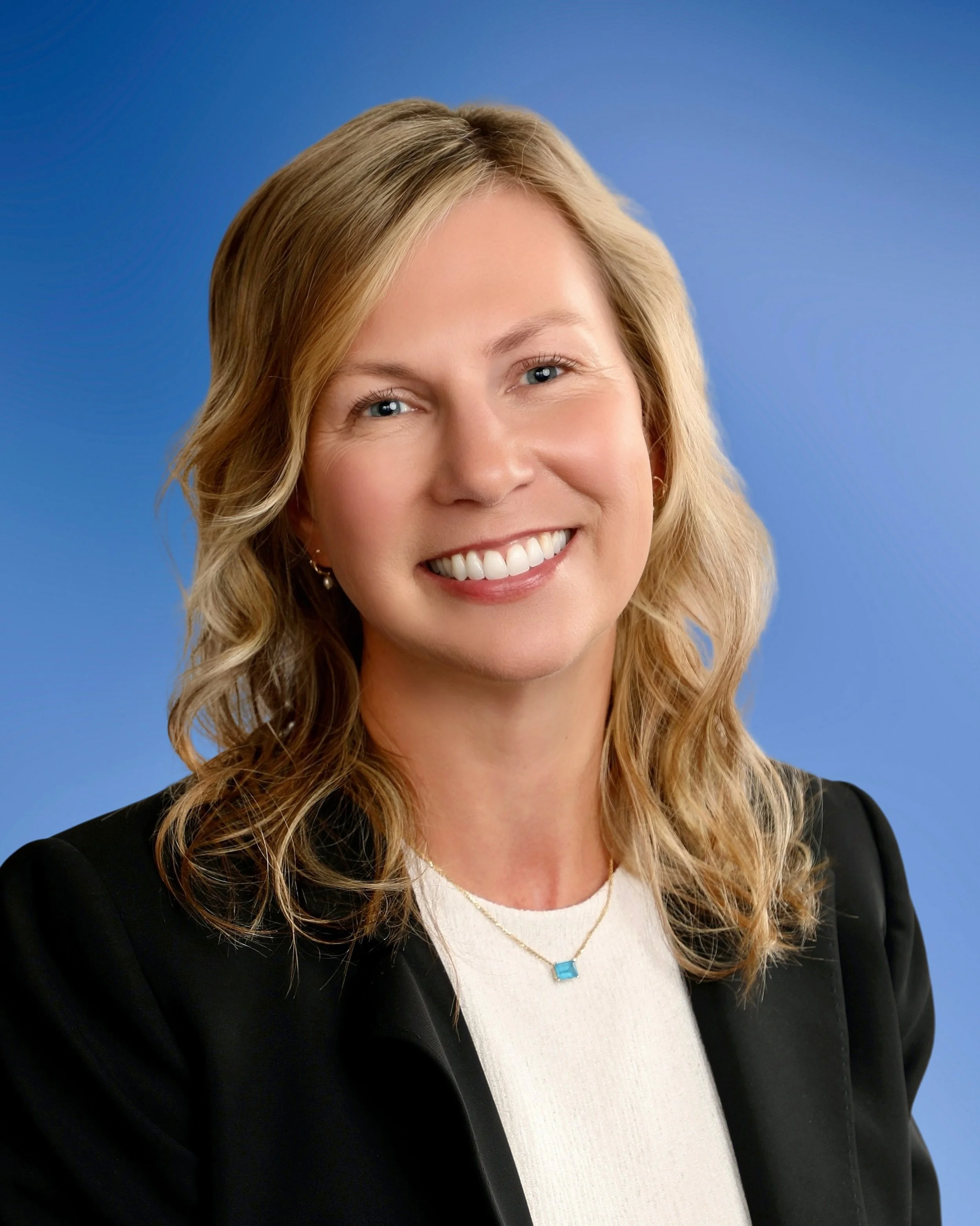 Close-up of a smiling woman with blonde hair, wearing a white top, black blazer, and a turquoise necklace, against a blue background.