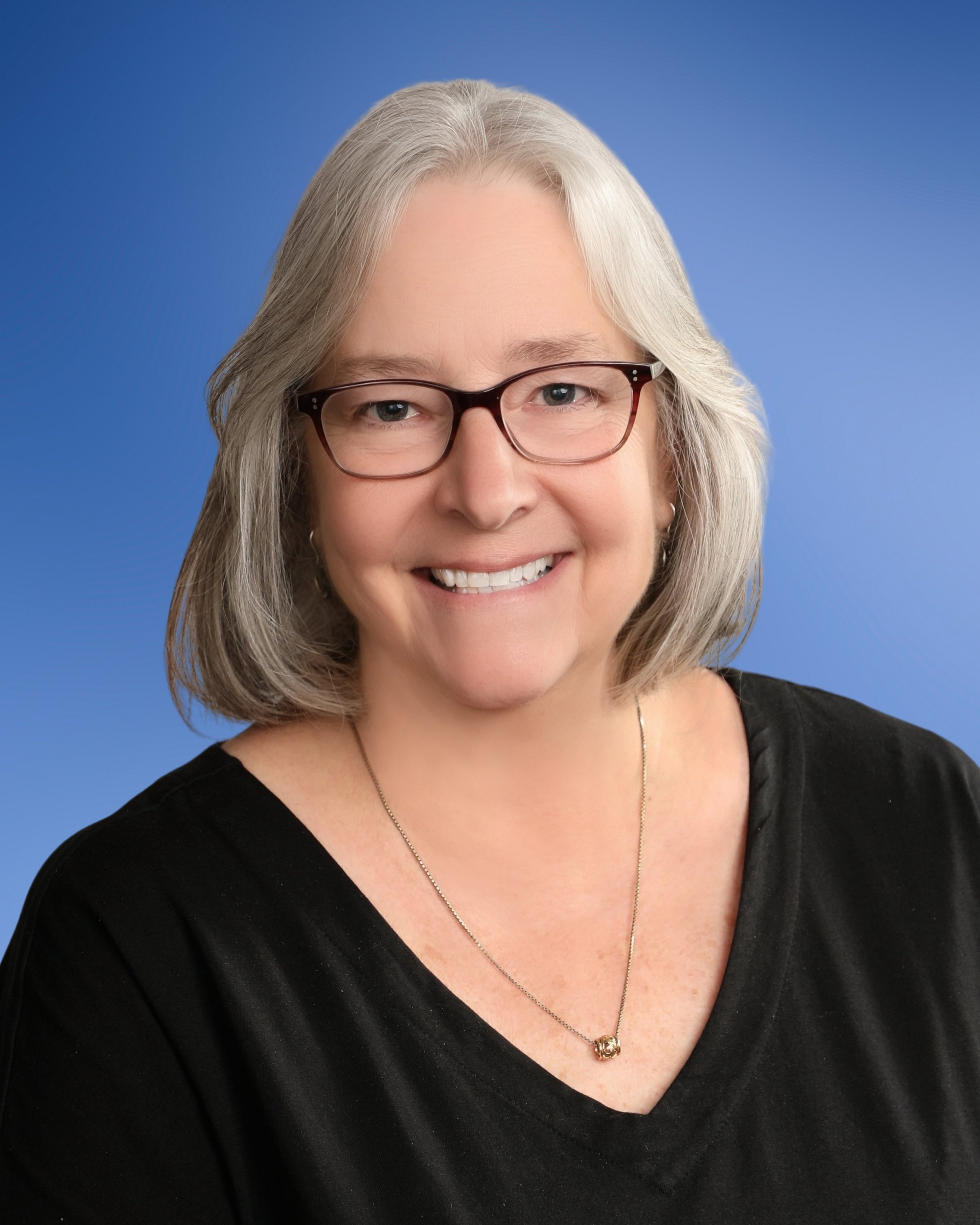 A smiling woman with shoulder-length gray hair wearing glasses, a black top, a necklace, and a pendant, against a plain blue background.