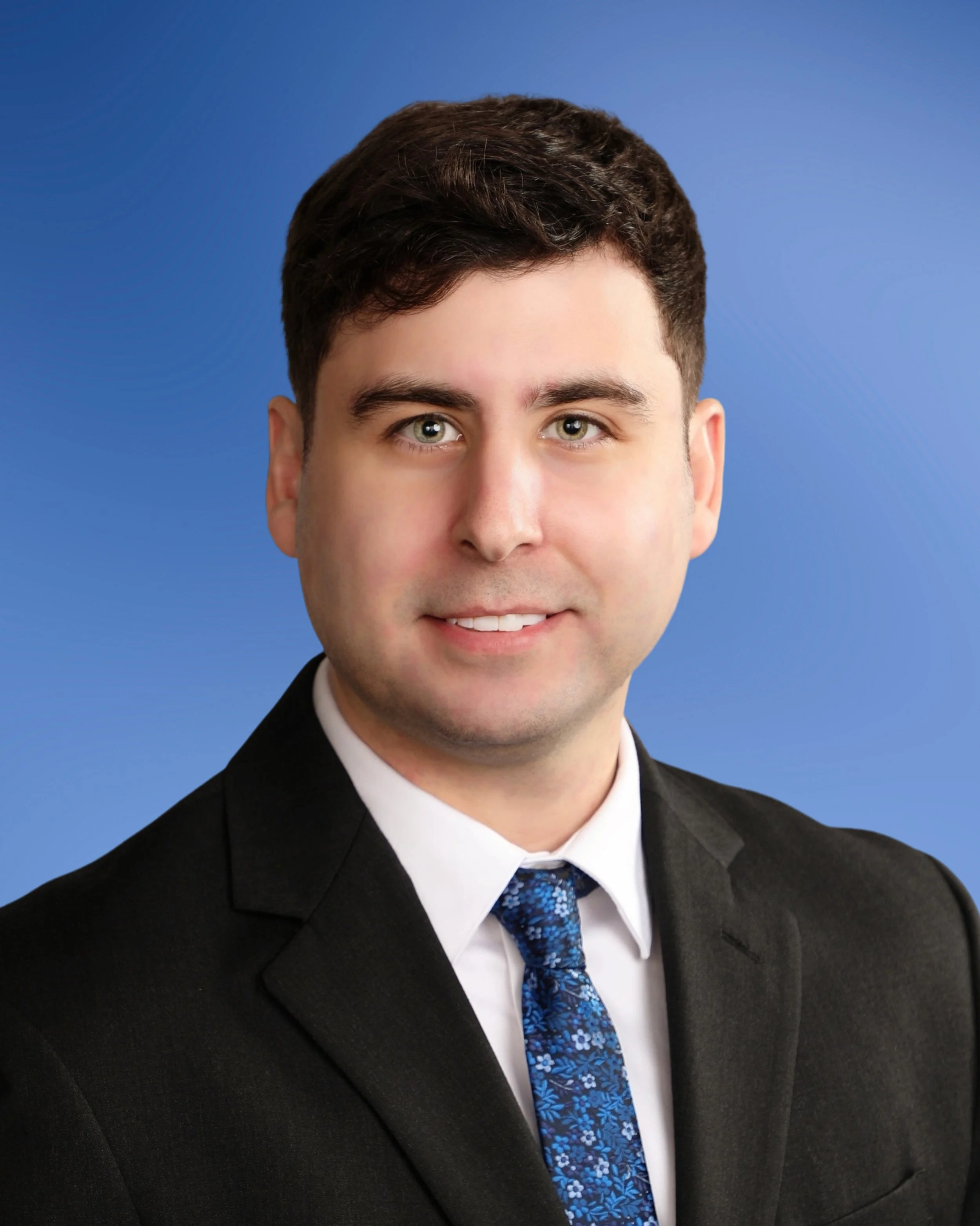 A young man wearing a black suit, white shirt, and blue patterned tie, smiling slightly against a blue background.