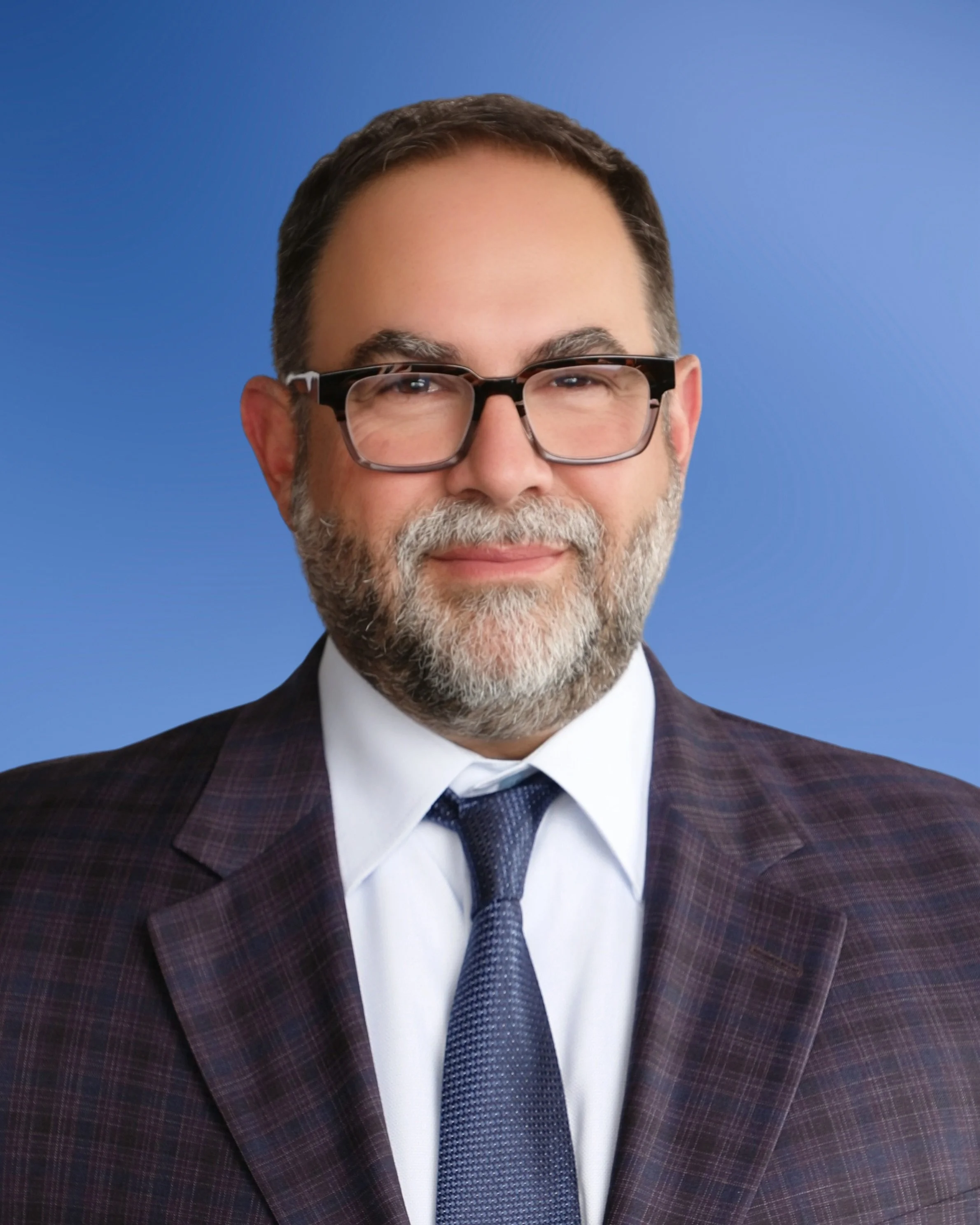 Close-up of a middle-aged man with glasses, smiling, dressed in a suit with a white shirt and navy tie, standing against a blue background.