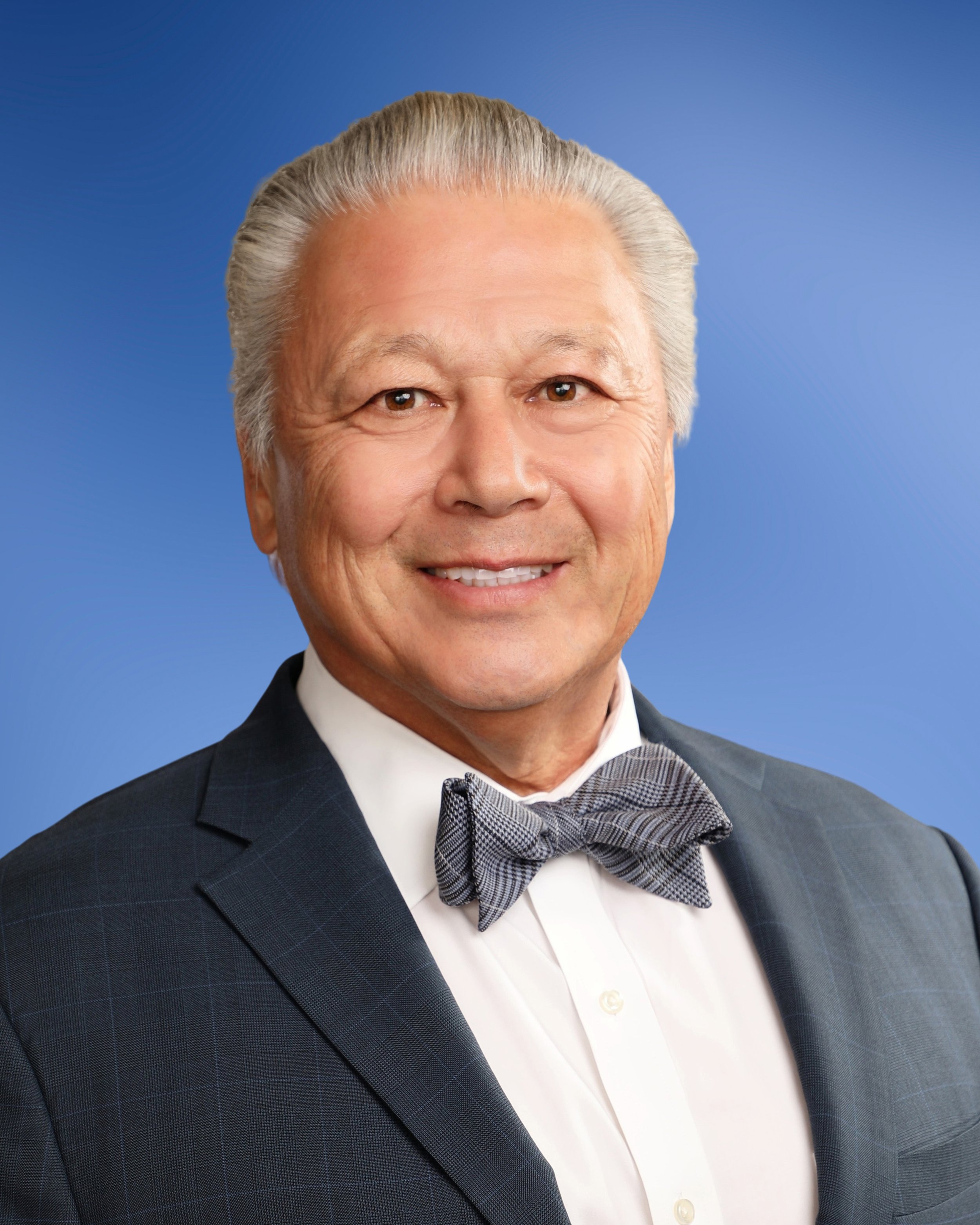 Close-up of a smiling older man with gray hair, wearing a tuxedo and bow tie, against a blue background.