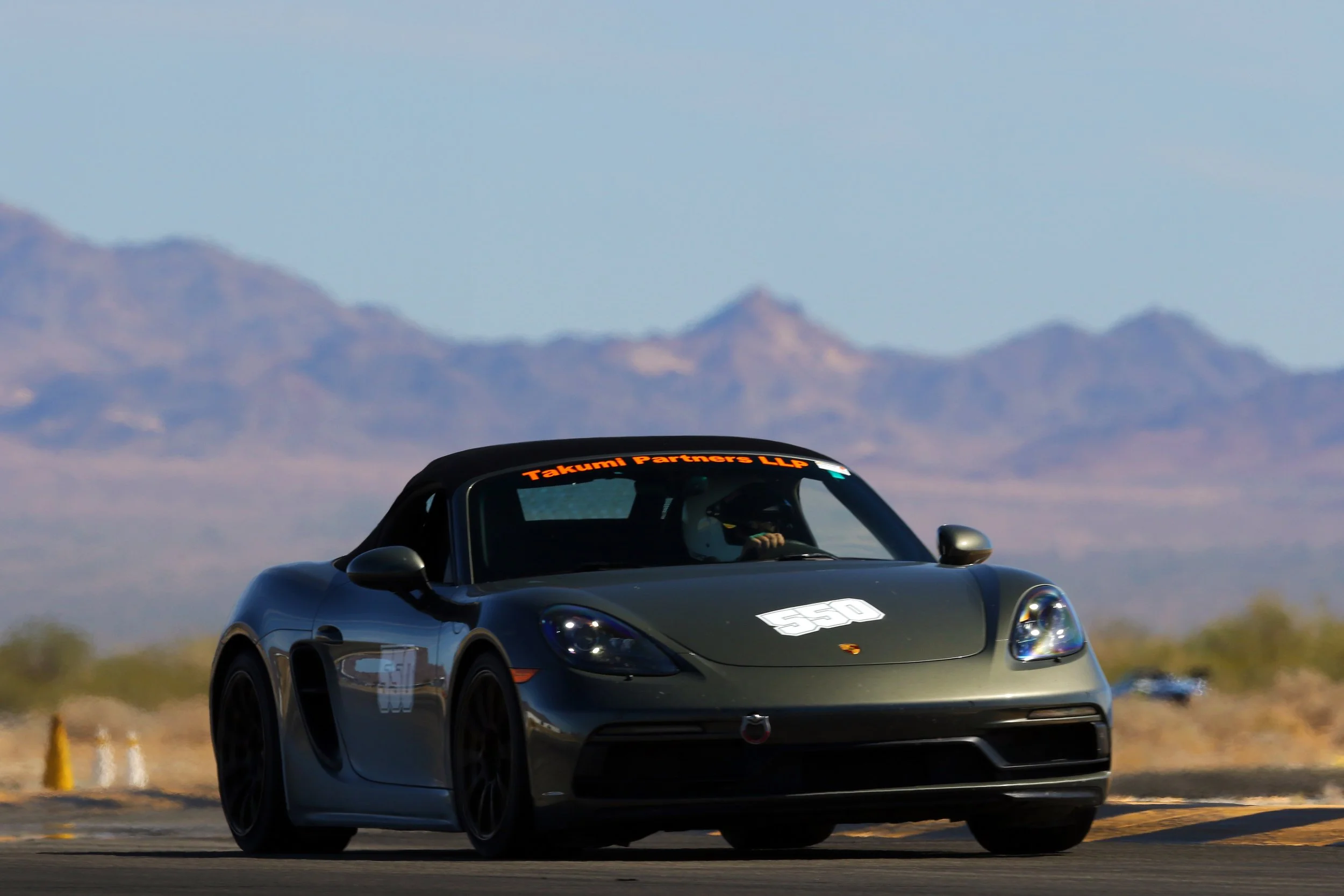 A black sports car with a racing number and the text 'Takumi Partners LLP' on the windshield, driving on a desert road with mountains in the background.