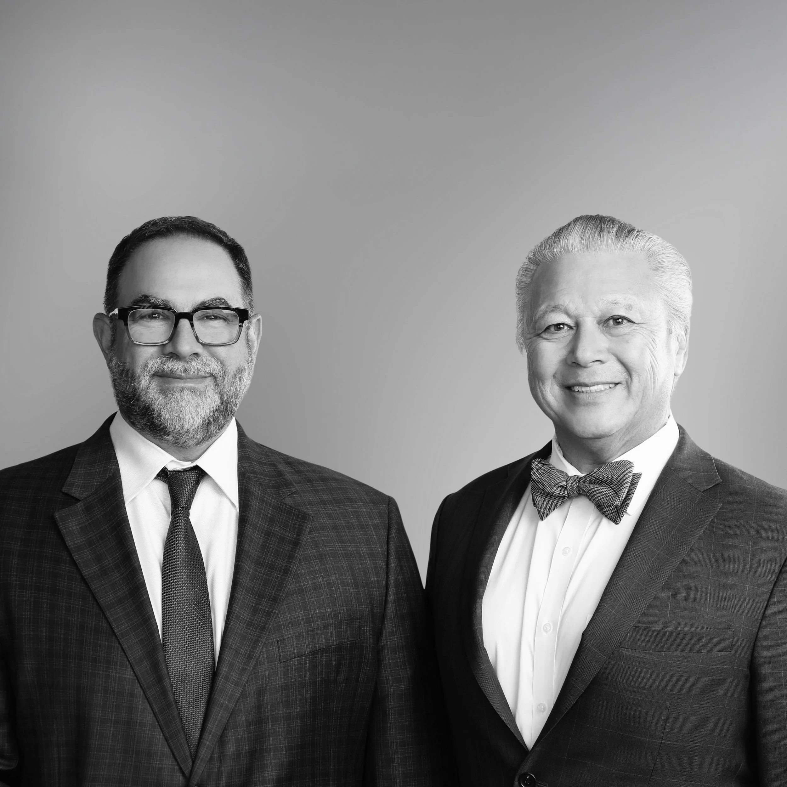 Close-up of two older men in formal suits, one with glasses and a beard, and the other with white hair, smiling against a plain background.