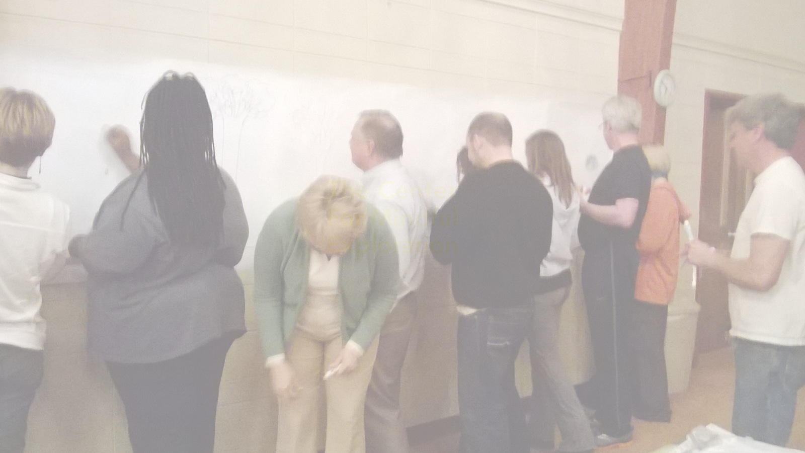 People standing in line at a whiteboard or glass board, some writing or drawing on it, in a classroom or conference room setting.