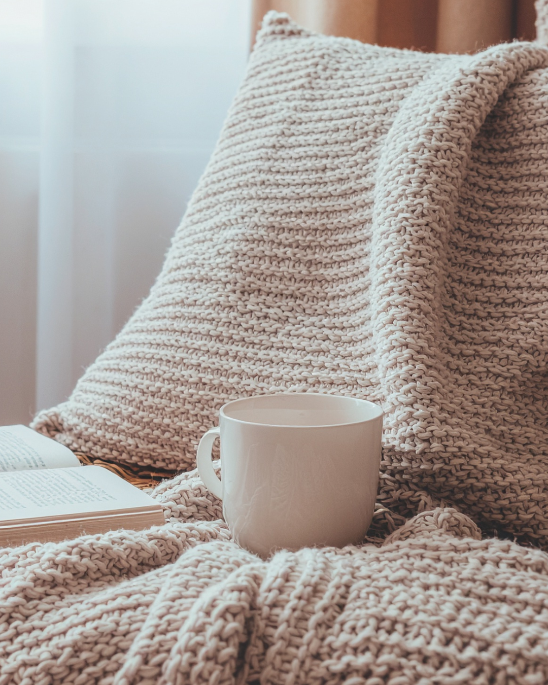 A cozy scene with a thick, knitted beige blanket draped over furniture, a white mug filled with a beverage, and an open book on a soft surface near a window with natural light.