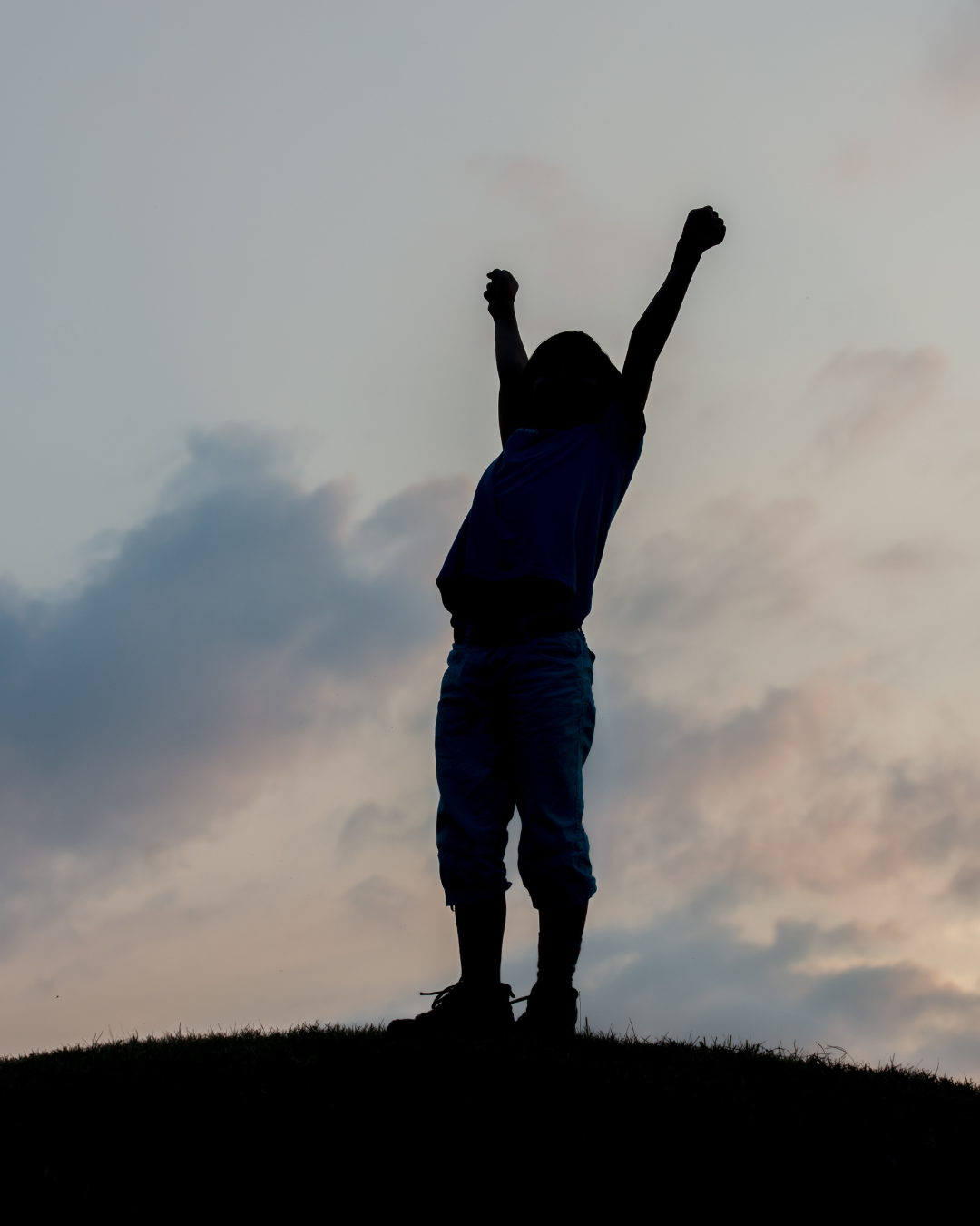 Silhouette of a child standing on a hill with arms raised in victory against a cloudy sky at sunset.