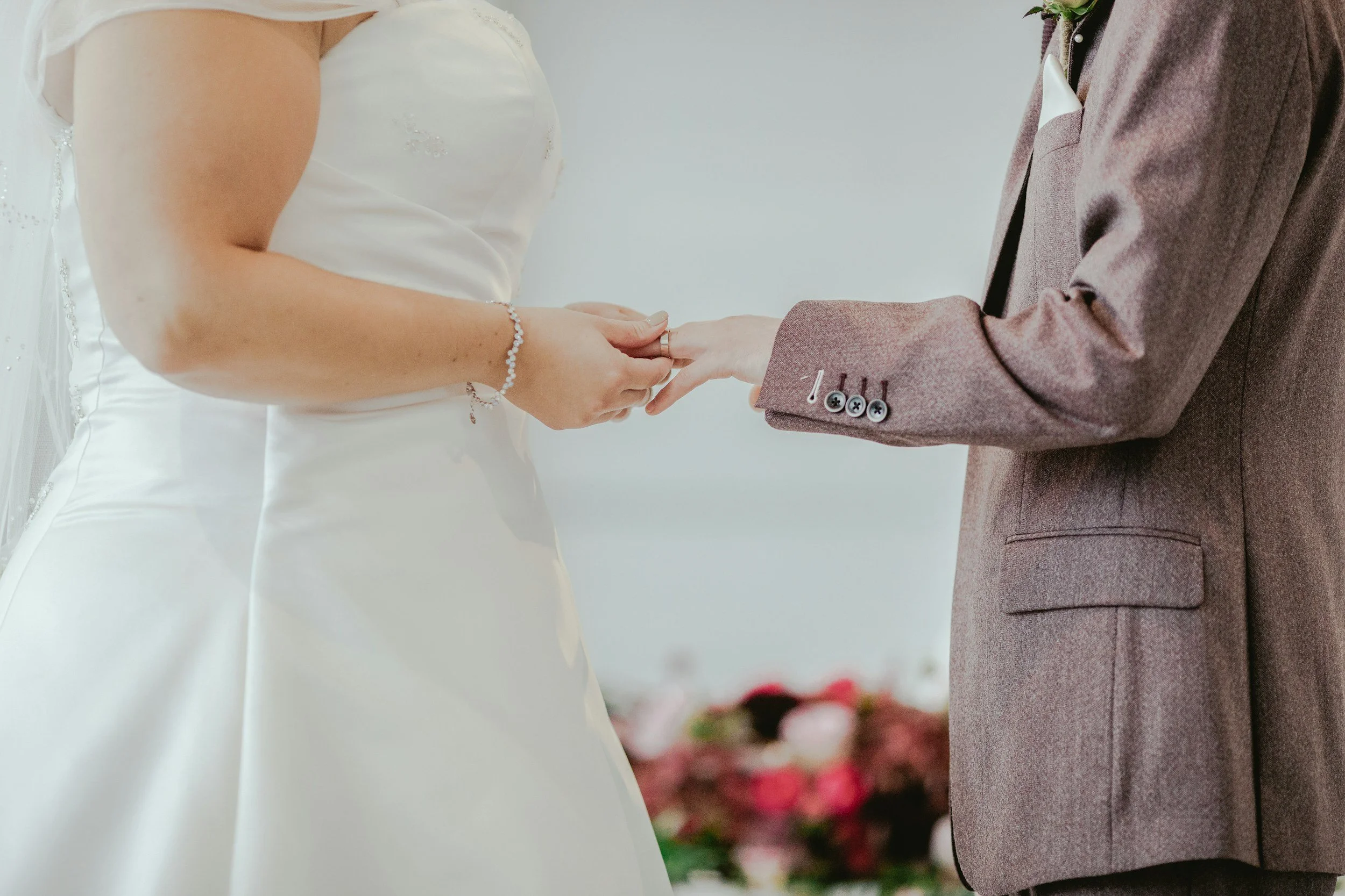 Ein Brautpaar hält sich bei einer Hochzeitszeremonie an den Händen, die Braut trägt ein weißes Kleid und Schmuck, der Bräutigam trägt einen braunen Anzug.