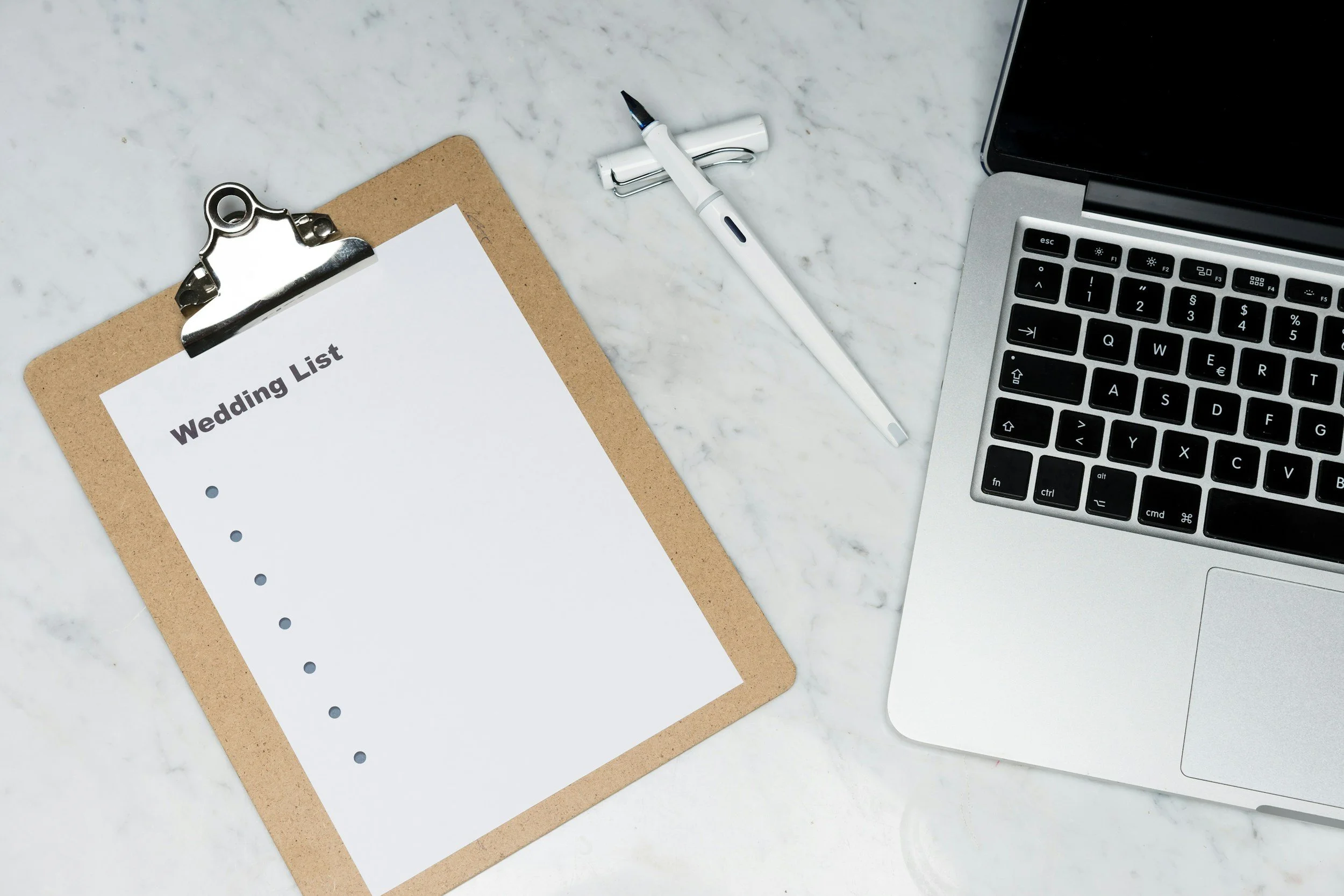 Arbeitsplatz mit Notizblock, Stift, Laptop und einem Dokument mit der Überschrift "Wedding List" auf einer Marmorfläche