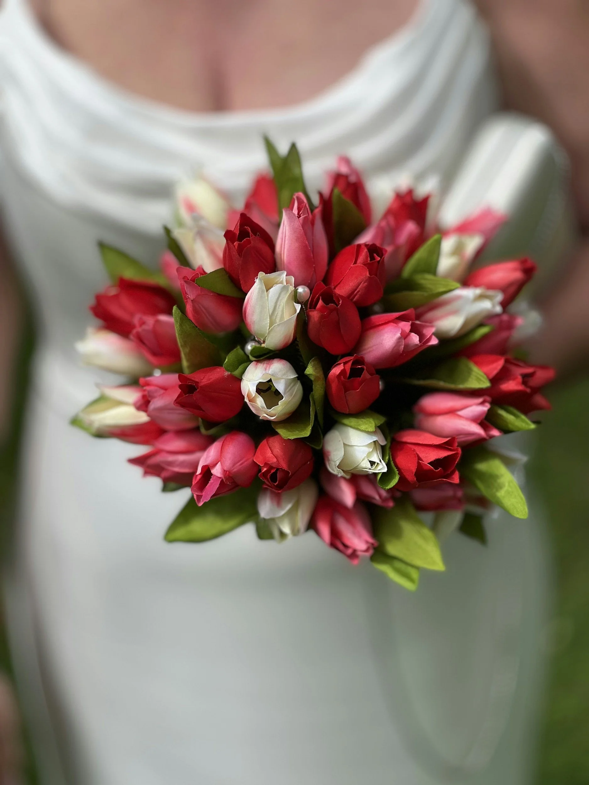 Ein Brautstrauß mit roten, weißen und rosa Tulpen, umgeben von grünen Blättern, wird vor einem weißen Kleid getragen.